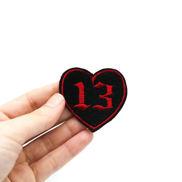 Embroidered black heart with a red outline and the number 13 in red in a gothic font. Held in a hand to show size 