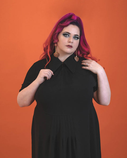 A model wearing a black 40s style short sleeved midi dress. elongated platter style collar and a narrow keyhole opening at the sternum with gathered detail and hook and eye closure. There is gathered pleating at the front of the full skirt. The plus size model is shown in close up holding the collar of the dress with both hands