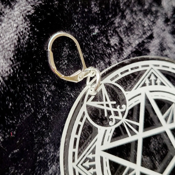 A pair of laser cut clear acrylic earrings in the shape of two circles etched with white symbols from a summoning circle. With silver plated latch hooks. Seen up close on a grey velvet background