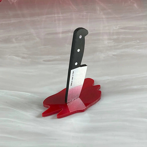 A ring holder in the shape of a knife made of wood and acrylic on a bright red base in the shape of a pool of blood. The knife is facing handle up.
