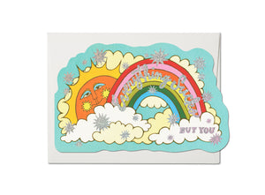 Die cut greeting card of a smiling sun surrounded by a rainbow and clouds. Lettering says “Everything Sucks But You” in script. Star details and lettering are silver holographic foil