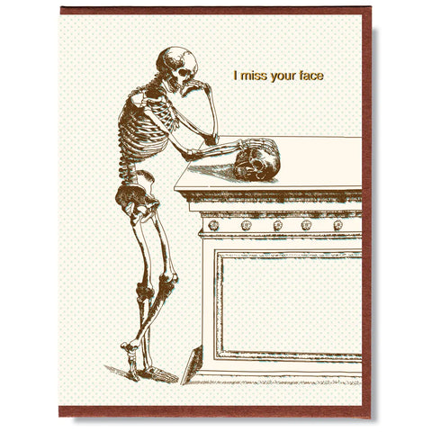 A rectangular note card of a skeleton standing against a ledge looking at a skull with the message “I miss your face” stamped in gold foil