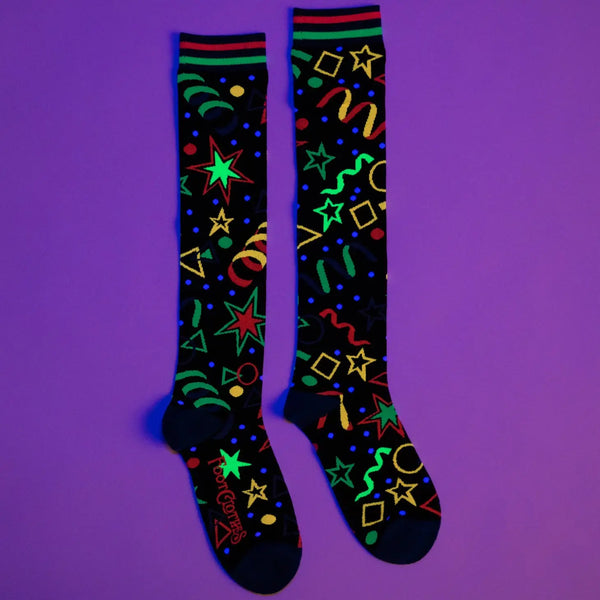 A pair of black knee socks with neon blue, green, and pink stripes at the cuff and blue toes & heels. There is a neon colored geometric pattern along the calf of each sock similar to an 80s bowling alley carpet. Shown under black light with the UV reactive fabri 