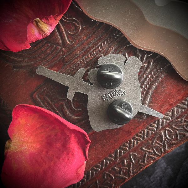 A pearly silver metal hard enamel pin with red detailing of an anatomical style heart being pierced by a 3D sculpted pointy dagger. Back of pin is shown on a wooden table surrounded by rose petals 