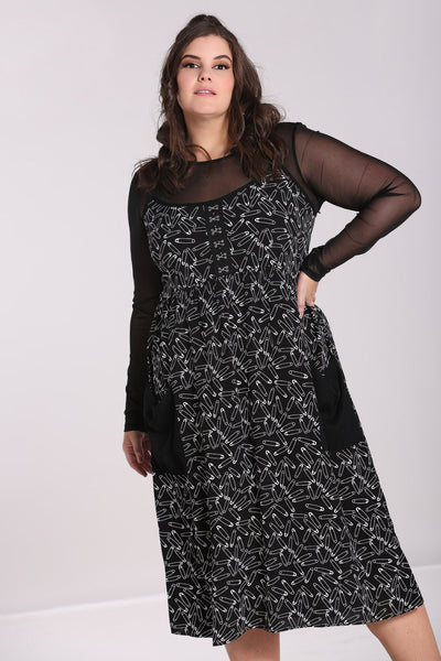 A plus size model wearing a black sleeveless sundress with an all-over white safety pin pattern. The dress has black spaghetti straps and silver metal hook and eye detail at the bodice. The skirt is midi length with two black patch pockets at the hips. The model is wearing a long sleeved mesh top underneath the dress