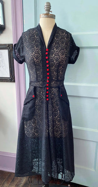 a black floral lace just past the knee dress. It has a v-neck shawl collar and cuffed short sleeves. There are bright red rose-shaped buttons running down the length of the bodice and on each patch pocket. Seen on a dress form from the front