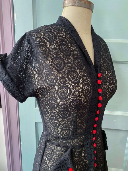 a black floral lace just past the knee dress. It has a v-neck shawl collar and cuffed short sleeves. There are bright red rose-shaped buttons running down the length of the bodice and on each patch pocket. Seen up close on a dress form from a three quarter angle