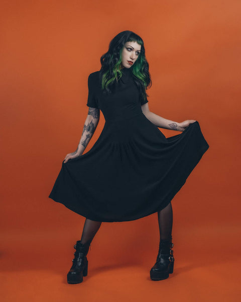 A model wearing a black 40s style short sleeved midi dress. elongated platter style collar and a narrow keyhole opening at the sternum with gathered detail and hook and eye closure. There is gathered pleating at the front of the full skirt. She is posing holding the skirt with both hands to illustrate its fullness 