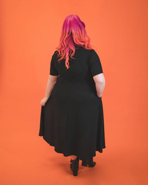 A model wearing a black 40s style short sleeved midi dress. elongated platter style collar and a narrow keyhole opening at the sternum with gathered detail and hook and eye closure. There is gathered pleating at the front of the full skirt. The plus size model is shown from behind to display the back of the dress