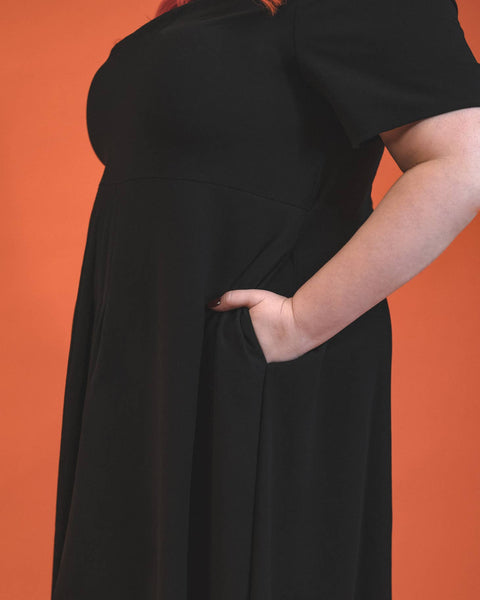 A model wearing a black 40s style short sleeved midi dress. elongated platter style collar and a narrow keyhole opening at the sternum with gathered detail and hook and eye closure. There is gathered pleating at the front of the full skirt. Zoomed in shot of the plus size model with their hand in one of the side seam pockets, shown from the side