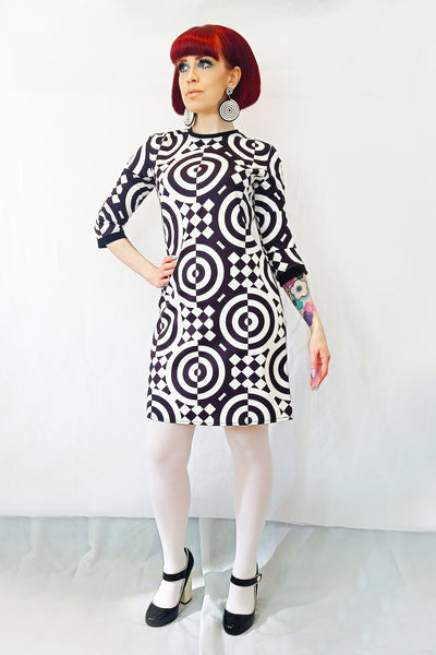 A full length shot of the model wearing a 3/4 sleeve mini dress with a black and white checkered and concentric circle op-art style pattern. It has a round neckline