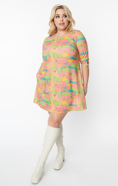 A plus-size model wearing a relaxed fit shift style mini dress with a jewel neckline and 3/4 sleeves with a keyhole detail. The pattern of the dress is a green, blue, pink, and orange 60s style illustration of a park with a frog wearing overalls playing guitar. The model has one hand at their side and another hand in one of the pockets of the dress