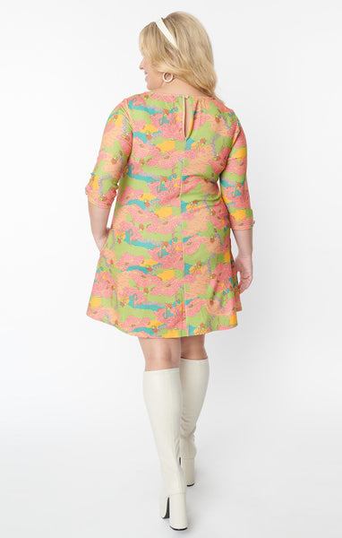 A plus-size model wearing a relaxed fit shift style mini dress with a jewel neckline and 3/4 sleeves with a keyhole detail. The pattern of the dress is a green, blue, pink, and orange 60s style illustration of a park with a frog wearing overalls playing guitar. The model is shown from behind to display the keyhole detail on the back of the dress