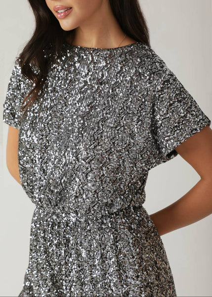 model wearing a short sleeved romper in silver sequin fabric with zigzag stripe pattern, round neckline, gathered elasticized waist, and deep keyhole detail at back. Shown from front in detail of sequin pattern