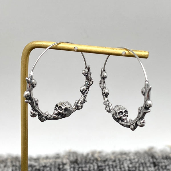 stainless steel infinity closure hoop earrings with skull and branch detail at the bottom of each hoop with tiny skulls growing out of each branch. Shown hanging