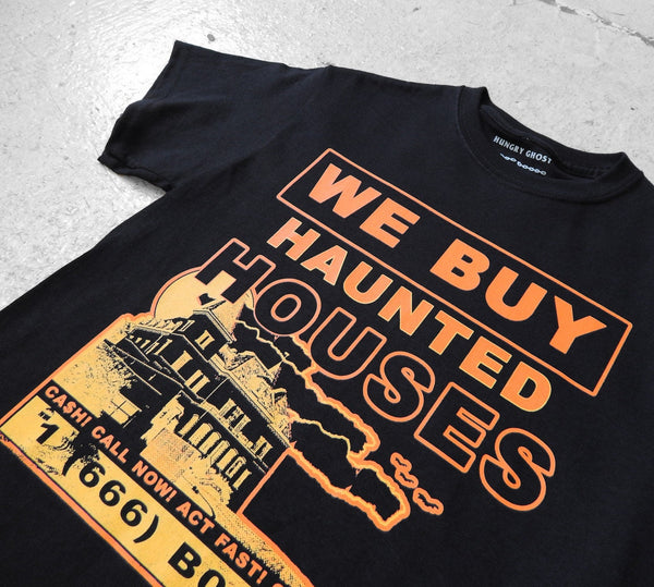 A black short-sleeve t-shirt with a vintage-sign style orange and yellow graphic that reads 'WE BUY HAUNTED HOUSES' above an illustration of a house and contact information: "CA$H! CALL NOW! ACT FAST! CASH FOR GHOSTS! 1 (666) B00-0000”. Shown in close up of print