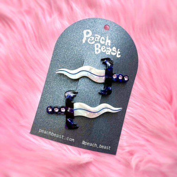 pair of acetate hair clips in the shape of small daggers with wavy iridescent blades and marbled blue and white handles with a row of three rhinestones in each. Shown on packaging card