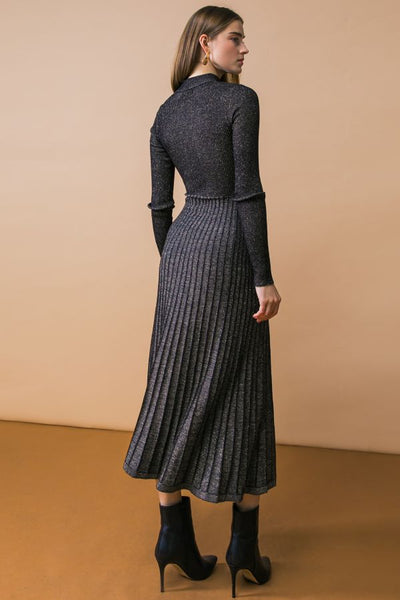 A model wearing a black and silver lurex knit dress with high neckline, long sleeves, fitted waist, and below the knee pleated skirt. Shown from back