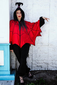 A model wearing a black fleece jumpsuit made to resemble Lydia Deetz from the cartoon Beetlejuice. It has a chunky black zip closure, red spiderweb style poncho, and matching black hood meant to resemble Lydia's hair in a ponytail. Shown with hood up