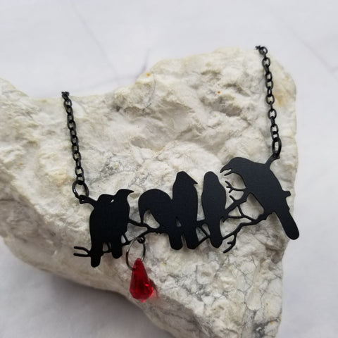 necklace with large black enameled metal pendant of six crows seen in silhouette on a branch with attached faceted red teardrop bead. On a 17" black enameled link chain