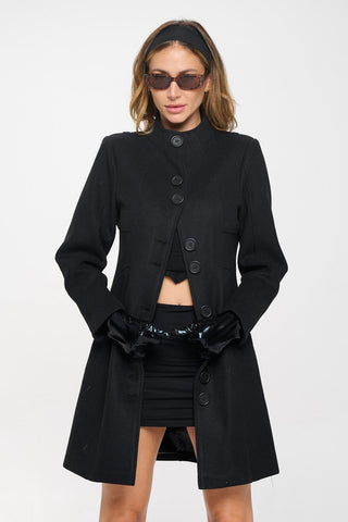 Model wearing a black faux wool single breasted knee length coat with funnel neckline, slash pockets, a banded waist, and black matte plastic buttons down the coat placed in sets of two. Shown from the front