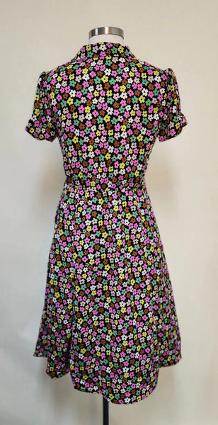 short sleeve dress in a black background magenta, green, brown, beige, and cream stylized flower print. It features short puffed shoulder sleeves with small gathered detail at the hem, darted bodice with a v-neck notched collar and black plastic button closure to the fitted waist, flared just-below-the-knee length skirt. shown back view on a dress form