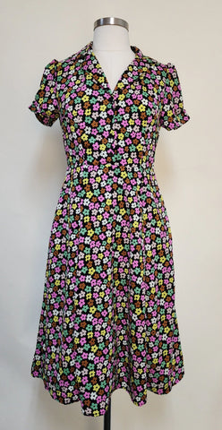 short sleeve dress in a black background magenta, green, brown, beige, and cream stylized flower print. It features short puffed shoulder sleeves with small gathered detail at the hem, darted bodice with a v-neck notched collar and black plastic button closure to the fitted waist, flared just-below-the-knee length skirt. shown on a dress form