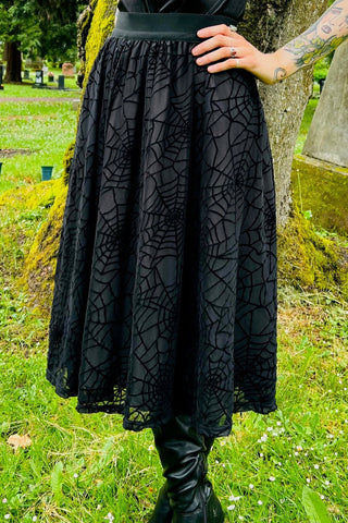 Model wearing a high waisted black below the knee length full skirt with a 2” wide elastic waistband, outer layer of sheer black mesh with a black flocked spiderweb design, and inner lining. Shown from the front