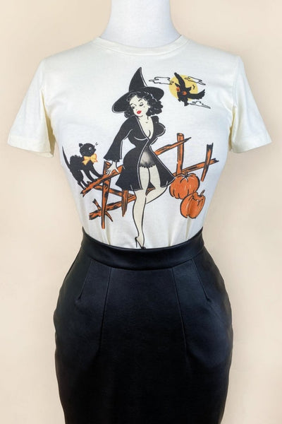 off-white womens cut t-shirt with illustrated graphic of pinup style woman wearing black negligee and witch hat resting on a wooden fence with hissing black cat, owl, and two pumpkins. Shown tucked into a leather skirt on a dress form