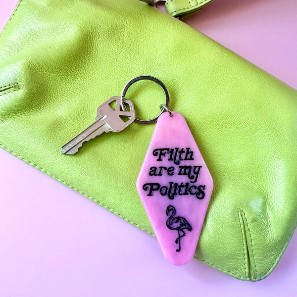 A pink acrylic hotel-style keychain with the text 'Filth are my Politics' and a flamingo graphic in black, attached to a metal key ring and displayed on a lime green leather clutch against a light pink background.