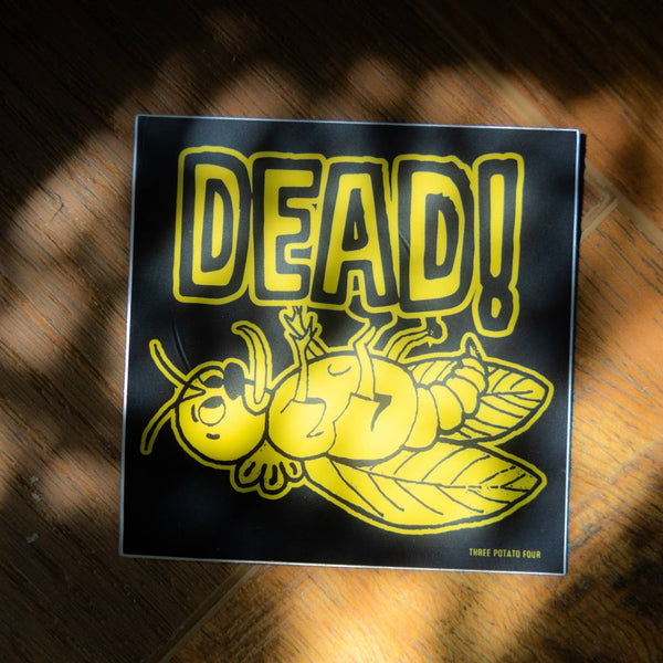 square vinyl sticker with matte finish and black background yellow illustration of a dead bug lying on its back with tongue sticking out and message "DEAD!" in matching yellow lettering. Shown in natural lighting on wooden background
