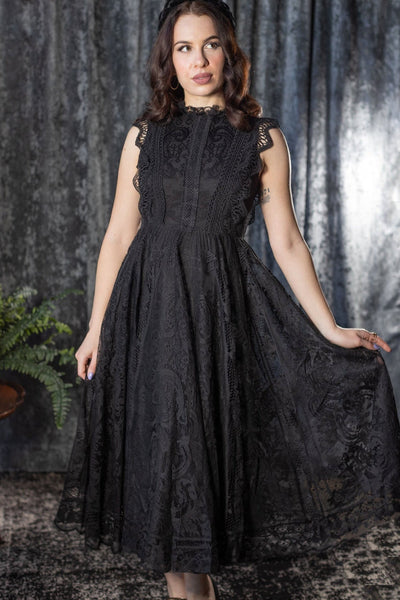 Model wearing a sleeveless midi dress with solid black shell and lace outer layer. It has a high neckline with ruffled lace collar, scalloped openwork lace at each arm opening, and a fitted bodice with three rows of geometric lace running vertically to the waist. It has a midi length lace skirt. Shown from front with skirt pulled taut