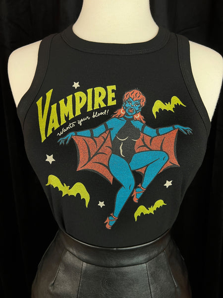 Cropped ribbed tank top with printed illustration of vampire woman with blue skin, red mullet, hoop earrings, a black bodysuit and red spiderweb patterned cape. Message written in yellow and white is "VAMPIRE wants your blood!" alongside yellow bats and white stars. Shown on a dress form in close up