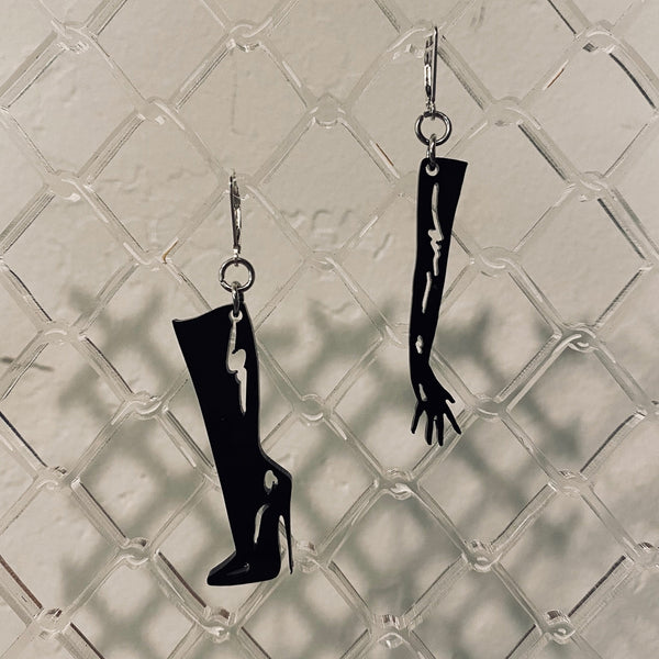 Black high-heeled boot and long glove-shaped earring set on a textured gray background