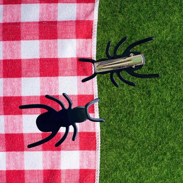 Black acetate ant-shaped hair clips on a red and white checkered cloth and green grass background