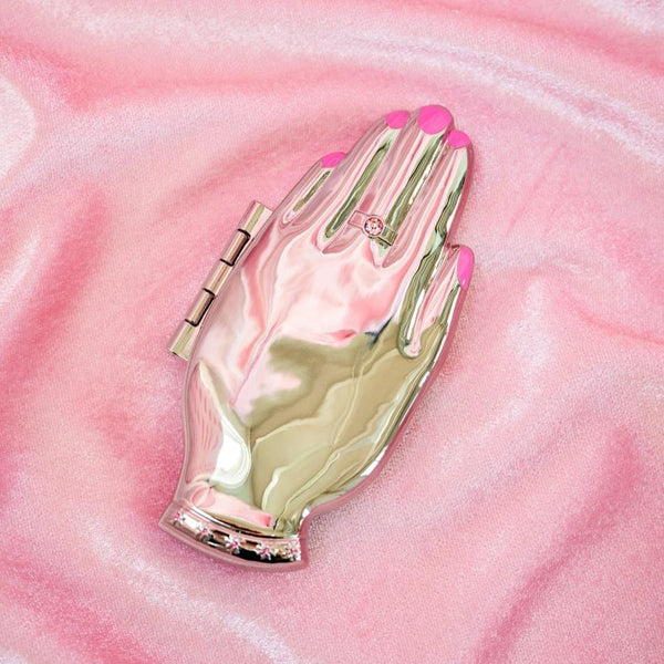 shiny finish silver metal hand-shaped compact mirror with bright pink enameled manicure and pink jeweled ring and bracelet detail. shown closed on pink satin background