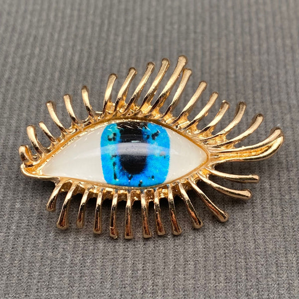 gold metal almond eye-shaped brooch with resin inset eyeball with blue iris and fanned out gold metal eyelashes. Shown from front