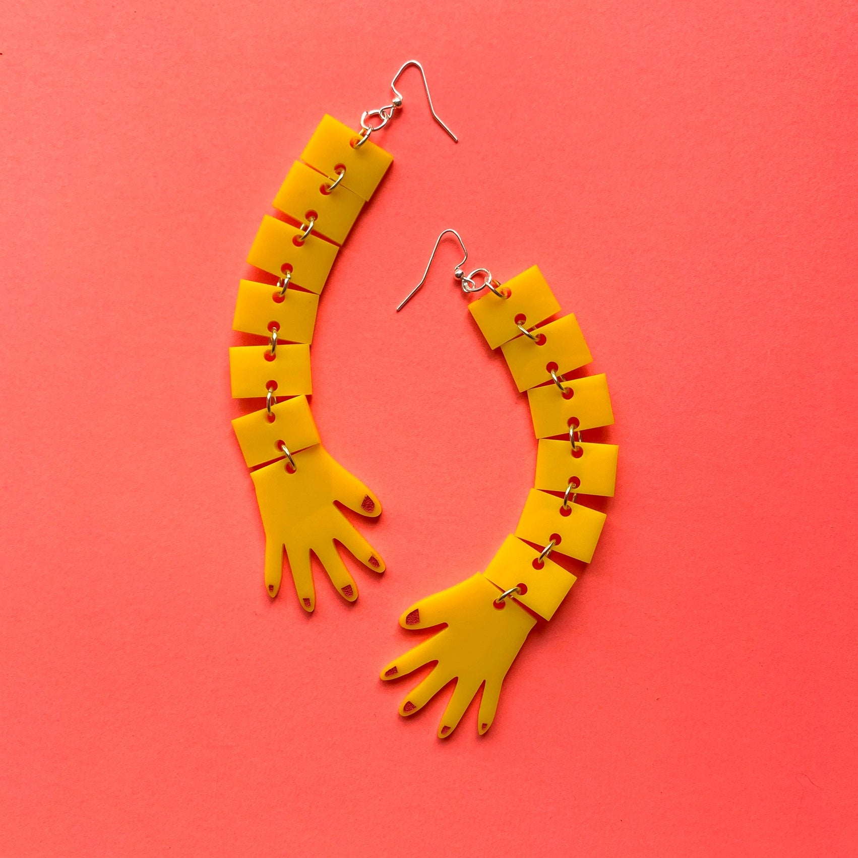 Bright yellow laser-cut acrylic dangle earrings in the shape of two long segmented arms and hands with red fingernails. On a bright pink background 