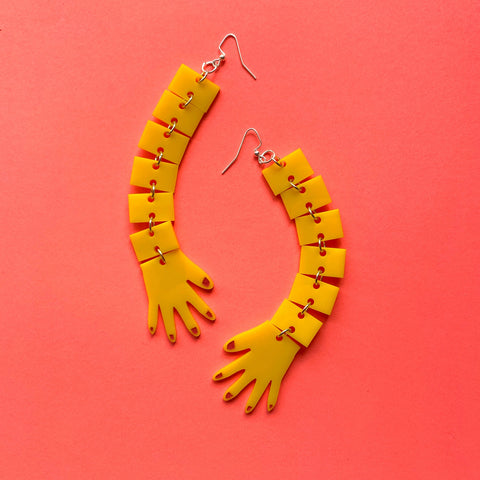 Bright yellow laser-cut acrylic dangle earrings in the shape of two long segmented arms and hands with red fingernails. On a bright pink background 