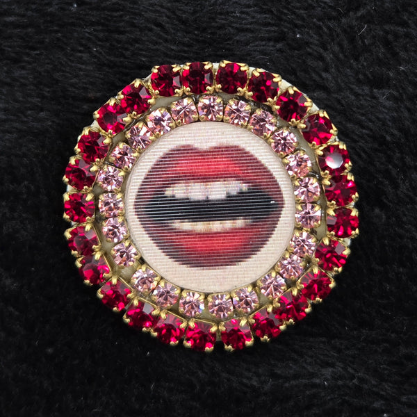 round brooch with lenticular red-lipped mouth design and rhinestone embellishments on a black background