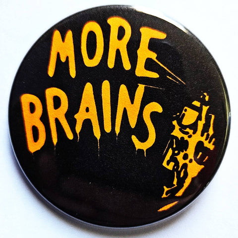 1.25" round pinback button with black background and message "MORE BRAINS" spray painted by decaying zombie hand, both in bright orange