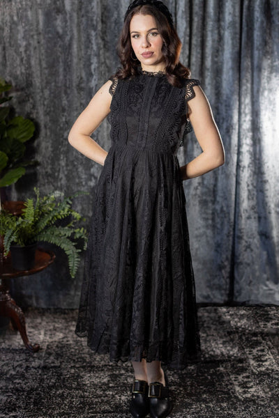 Model wearing a sleeveless midi dress with solid black shell and lace outer layer. It has a high neckline with ruffled lace collar, scalloped openwork lace at each arm opening, and a fitted bodice with three rows of geometric lace running vertically to the waist. It has a midi length lace skirt. Shown from front