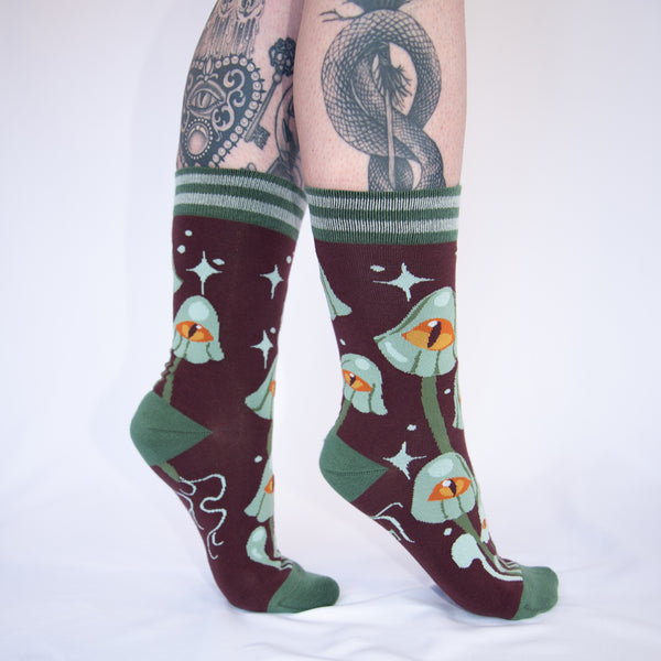 Unisex crew socks with an all-over pattern of green-grey mushrooms each with a large yellow and orange eye on their caps. On a purple-ish brown background surrounded by matching grey starbursts. Striped green and grey cuffs and solid green toes and heels. Seen worn by a model