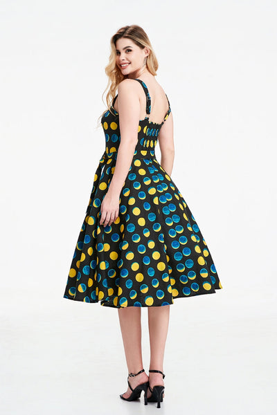 A model wearing a sleeveless black background yellow and blue moon phase print dress with a fitted sweetheart neckline princess seamed bodice with adjustable straps, removable self sash belt, swingy gathered full circle just below the knee length skirt. Shown from back