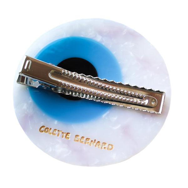 eyeball shaped hair clip with a white marbled white, red carved bloodshot details, a bright light blue iris, and a black pupil in the center, on a white background. Shown from back, displaying silver metal alligator style clip and gold branding