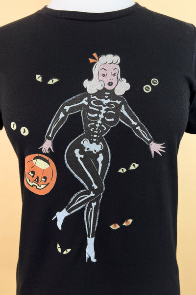 black short sleeve t shirt with a printed illustration of a pinup style woman wearing a skeleton costume holding a pumpkin basket surrounded by pairs of monster eyes. Seen on a dress form in closeup