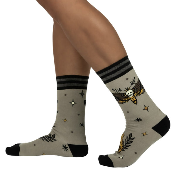pair of unisex cotton blend greenish beige background crew socks with pattern of brown and yellow death's head moth and caterpillar alongside tiny black and yellow stars. Shown worn by model from side