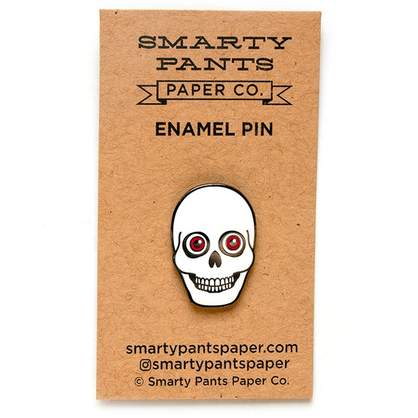 Black metal enamel pin of a white smiling skull with large red eyes shown on backing card