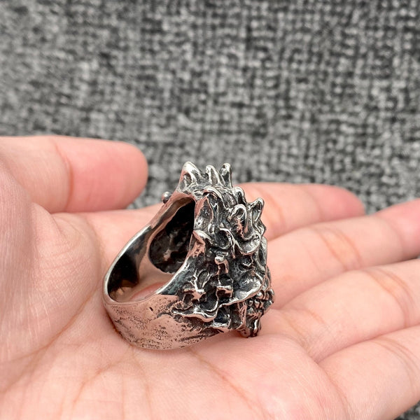 A stainless steel ring with antiqued detail in the shape of a demonic skull with horn-like features and grinning. Shown from the side