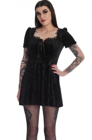 Model wearing a black stretch velvet mini dress with an all over embossed damask pattern of chandeliers, roses, candles, and crescent moons. It has lace trim at the bust and shoulders of the square neckline and a lace up notched detail at the bust. The skirt hits above the knee. Shown from the front